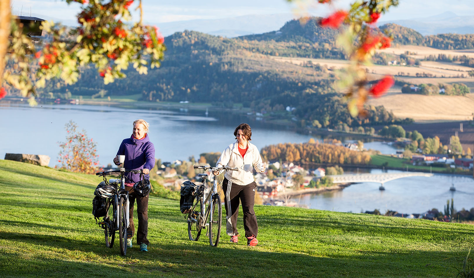  Two women with bikes om grass with landscape in the background. 