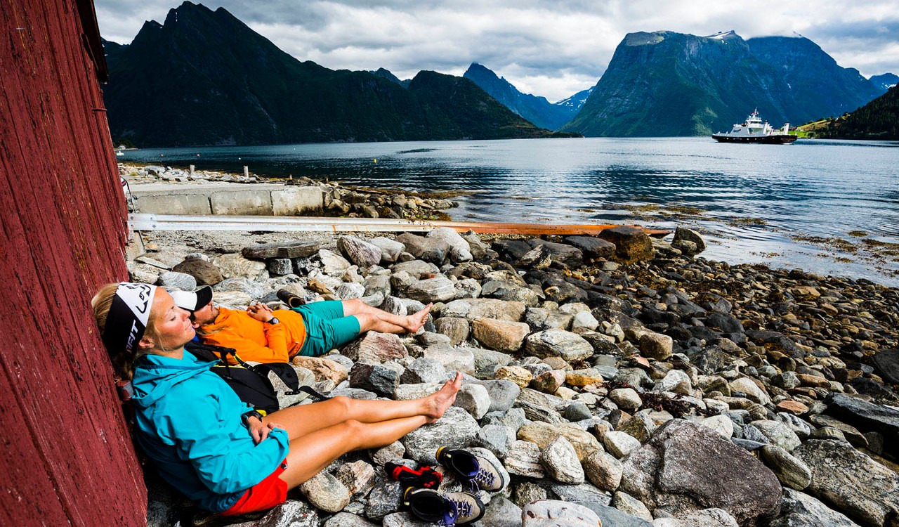 Turgåere slapper av i naustveggen med Hjørundfjorden foran seg på Sunnmøre, Vestlandet. Hikers relaxing on the stony beach of Hjørundfjorden, Sunnmøre, Vestlandet.