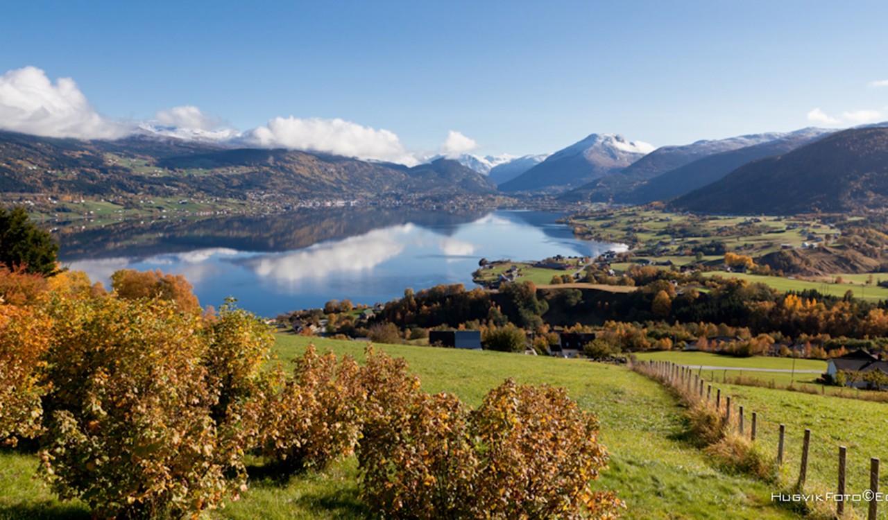 Utsikt over Gloppen med speilblankt vann, klar himmel, marker og fjell. View over Gloppen with water, blue sky and distant mountains.