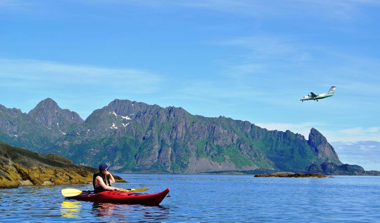 Mann i kajakk ser på Widerøe-fly som går inn for landing i Nord-Norge en sommerdag. Man in kayak watching a Wideroe plane preparing for landing in Northern Norway om a clear summer day. 