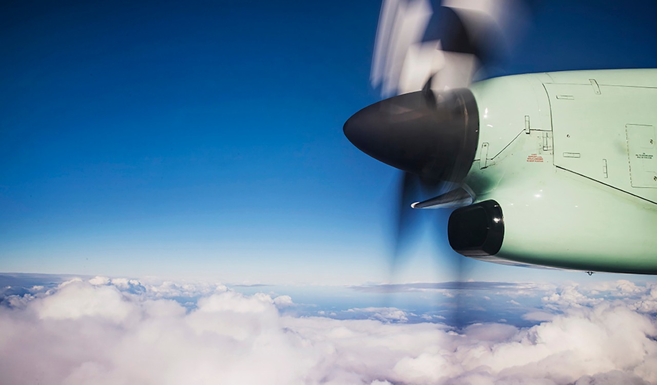 Propell sett fra flyvinduet med blå himmel og skydekke under. Propeller seen from the window of the plane with blue sky and white clouds beneath. 