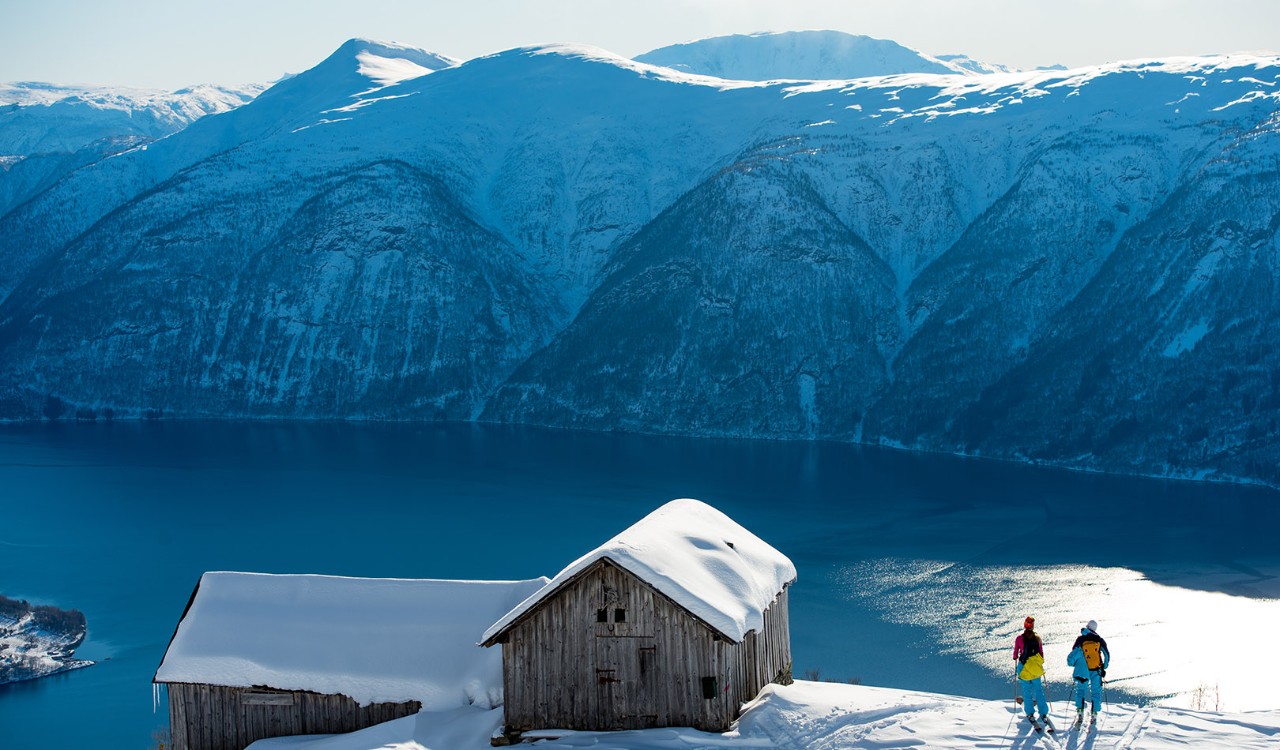 Skiløpere står ved siden av en låve på fjellet og ser utover fjord og fjell. Skiers standing next to an old barn watching mountains and fjords,.